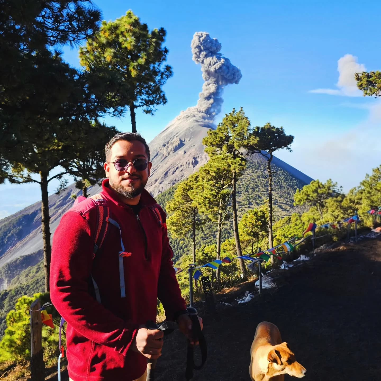 Ashish on the Acatenango trail in a red jacket, with a dog nearby and an eruption plume behind him.
