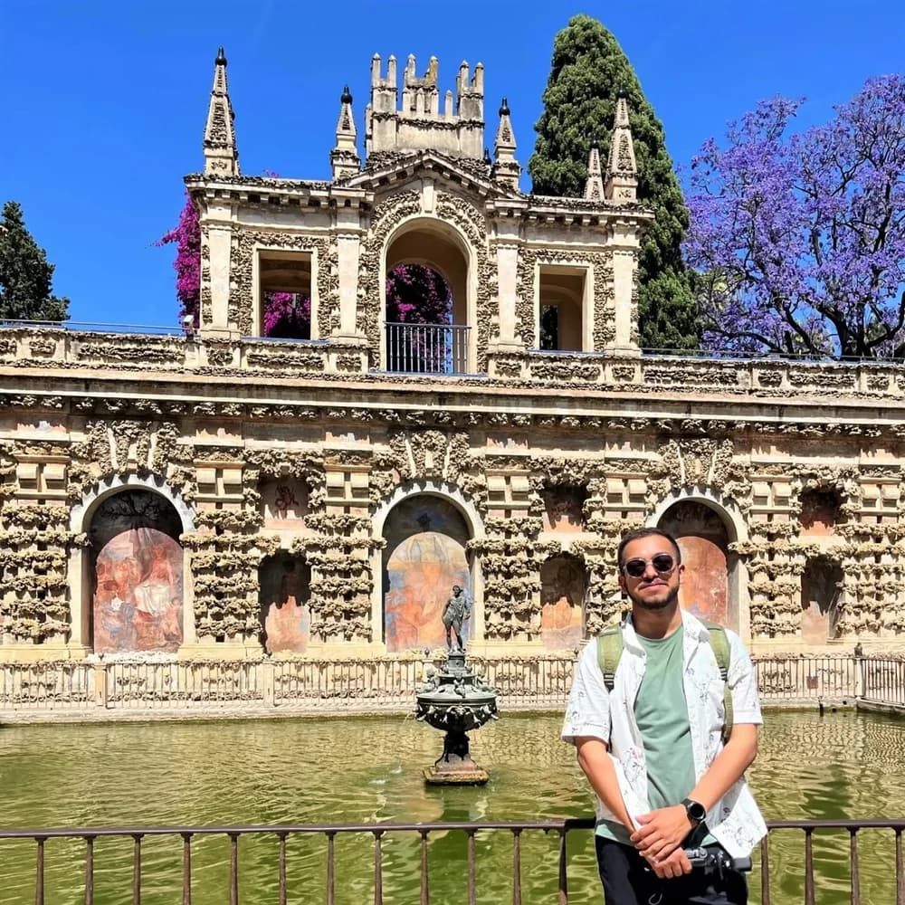 Ashish beside the ornamental pond in the Alcázar gardens in Seville, beneath an ornate stone gallery and a blue sky.
