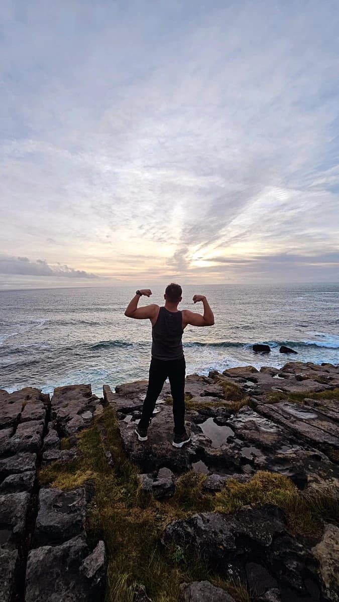 Ashish standing on a rocky shore with his back to the camera and arms raised, facing the Atlantic beneath a streaked evening sky.