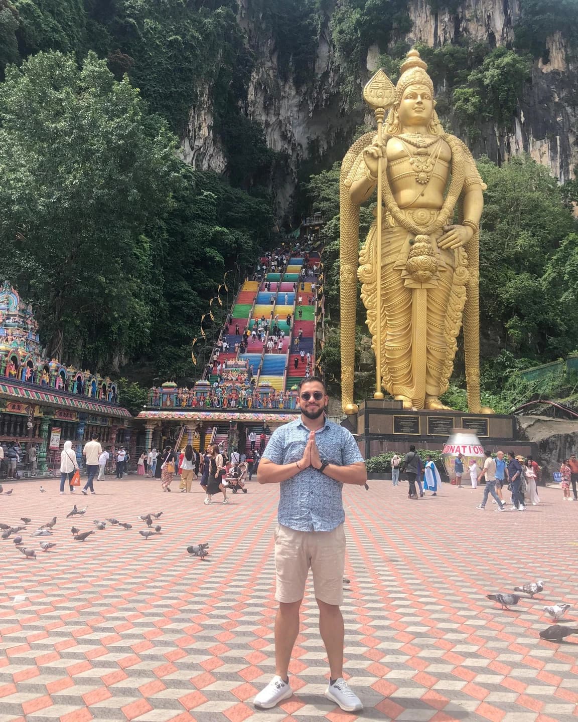 Ashish with his palms together at Batu Caves, with the golden Murugan statue, colourful steps and limestone cliffs behind him.