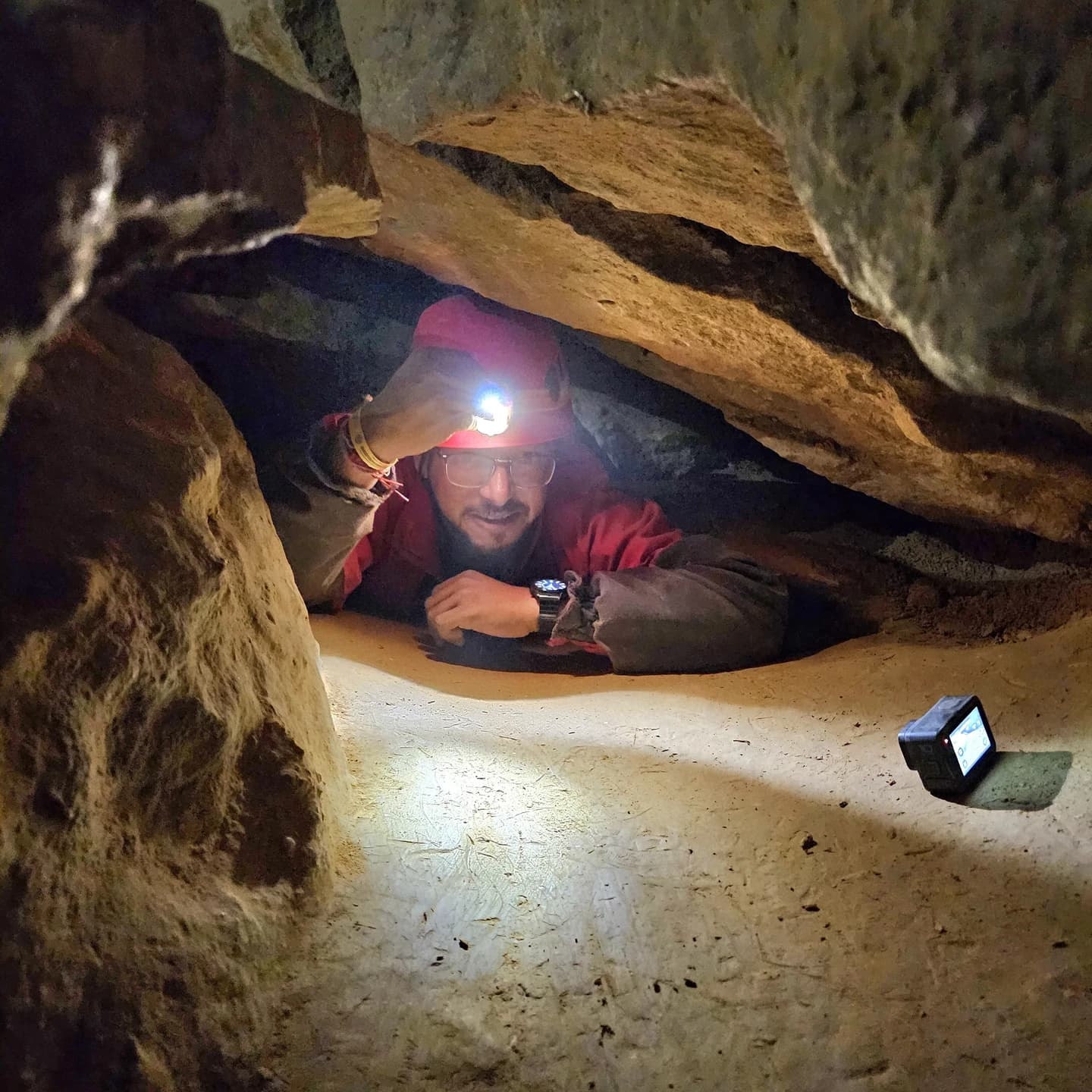 Ashish in red caving overalls and a headlamp, crawling through a narrow gap between limestone rocks.