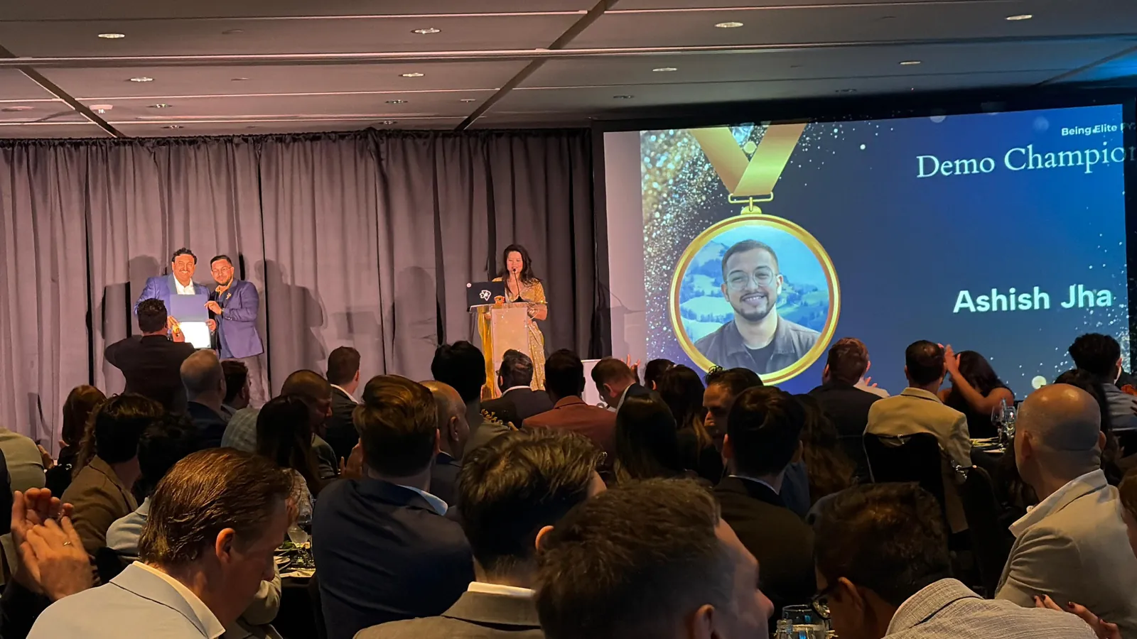 The DevRev SKO award presentation, with Ashish’s photograph and Demo Champion on the screen behind the presenters.