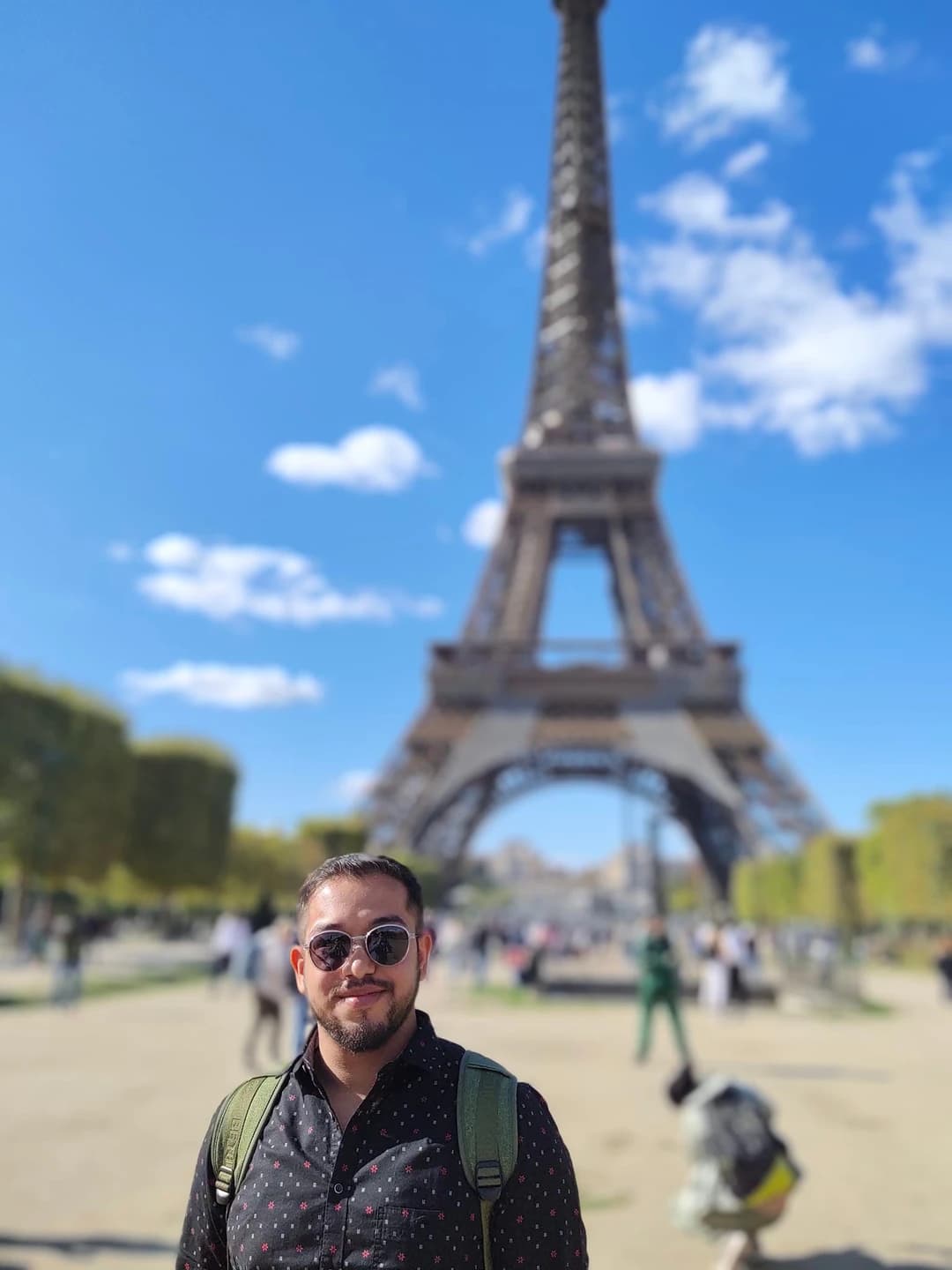 Ashish wearing sunglasses and a backpack, with the Eiffel Tower behind him against a blue sky.