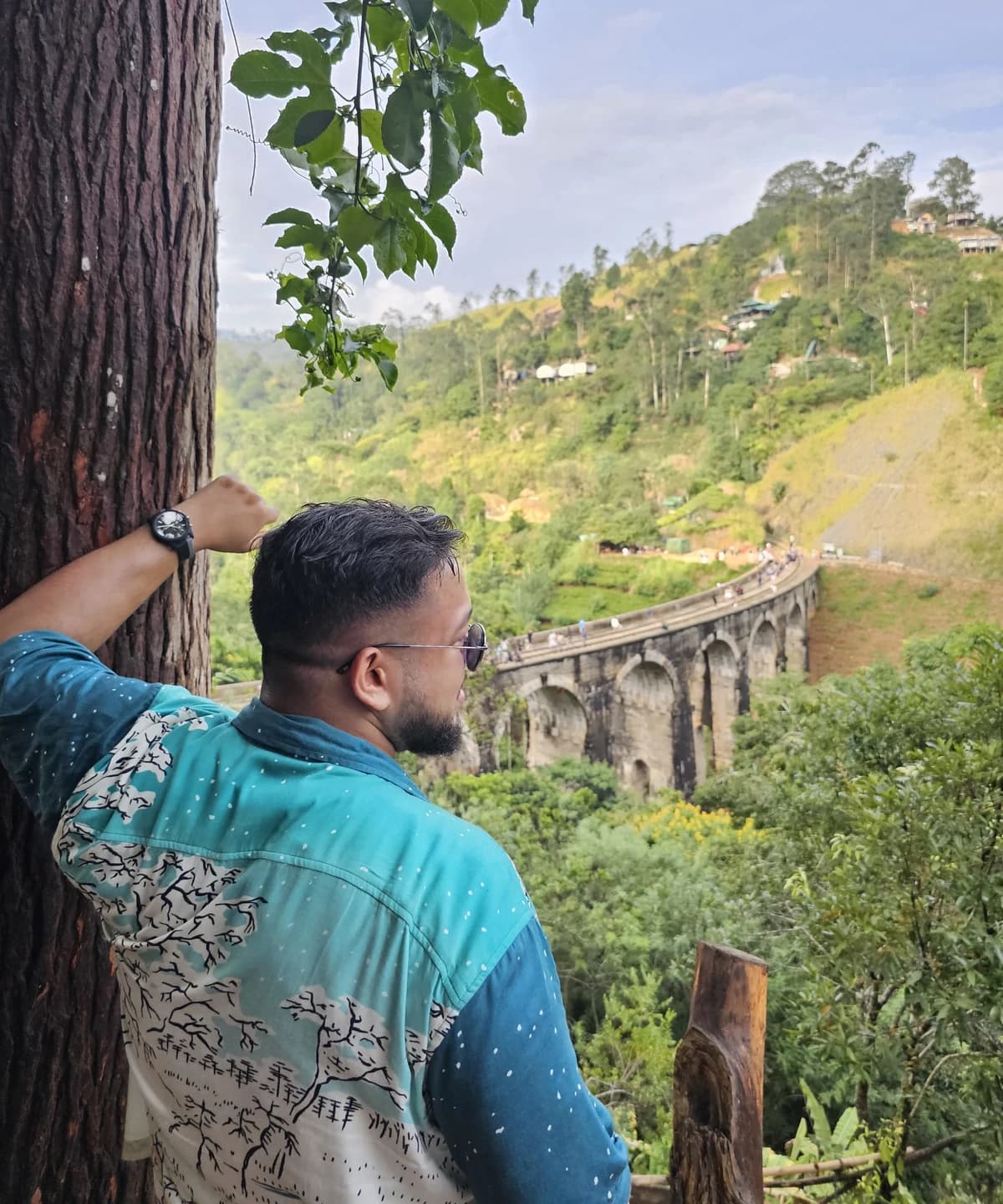 Ashish leaning against a tree, looking across the green hillside towards the stone arches of Nine Arches Bridge.
