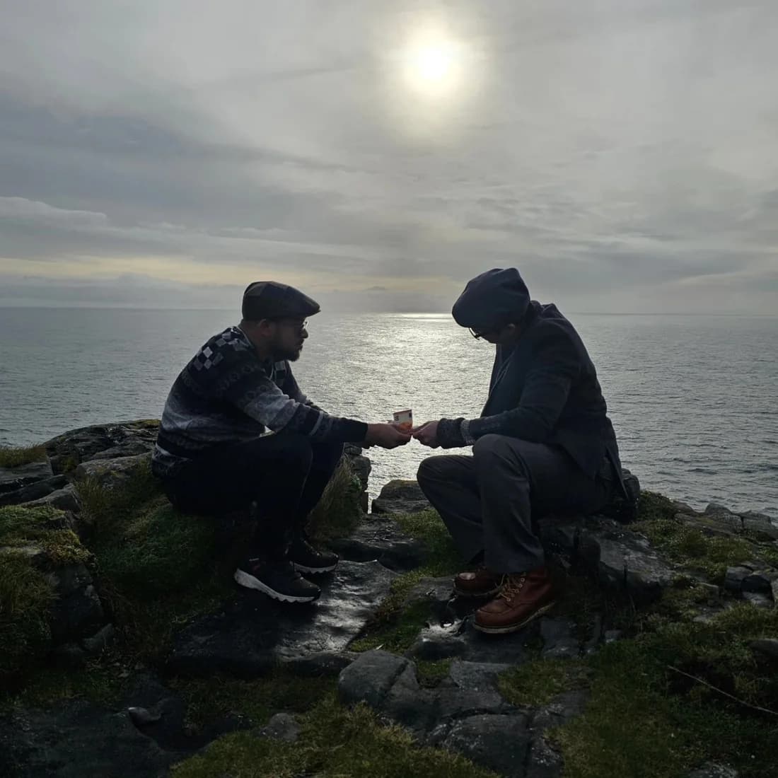 Ashish and a companion sitting on coastal rocks in Ireland, reaching toward each other, with the sea and a hazy sun behind them.