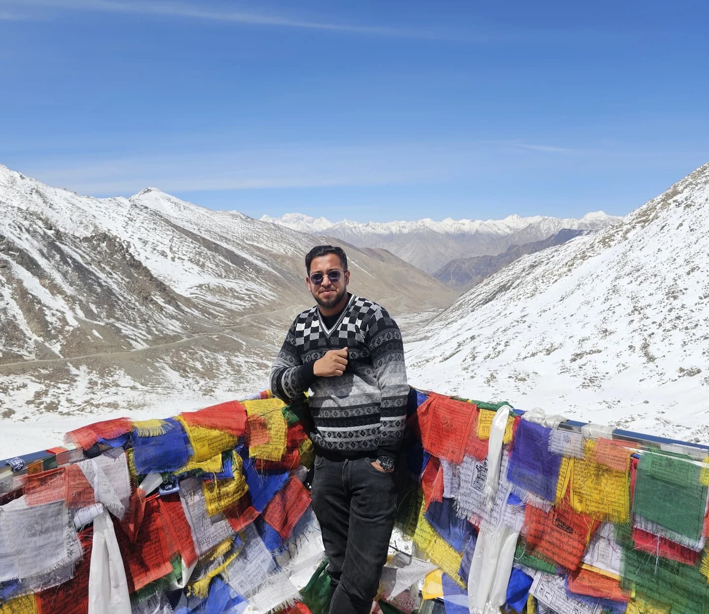 Ashish standing between colourful prayer flags, with a snow-covered mountain valley behind him.