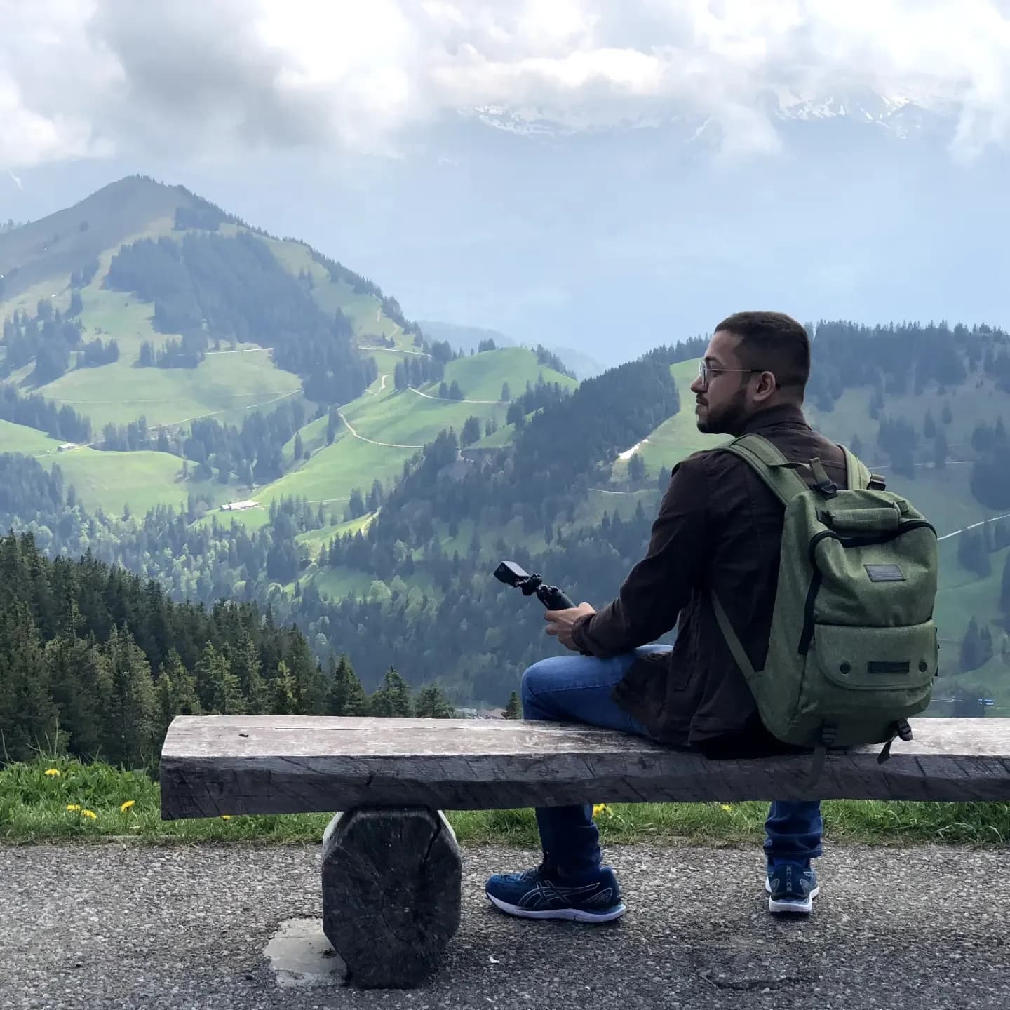 Ashish sitting on a bench with his backpack, looking across green mountains at Mount Rigi.