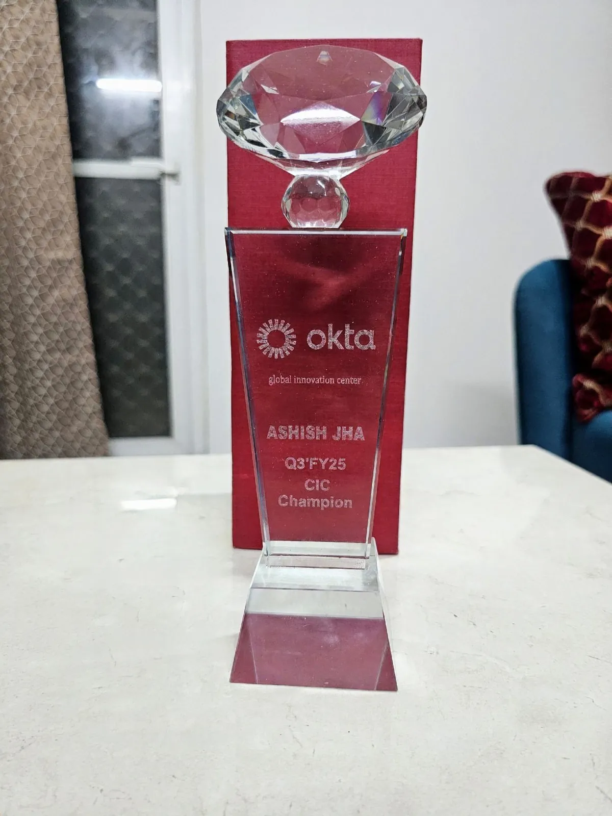 A red-and-clear Okta trophy engraved Q3 FY25 CIC Champion and Ashish Jha.