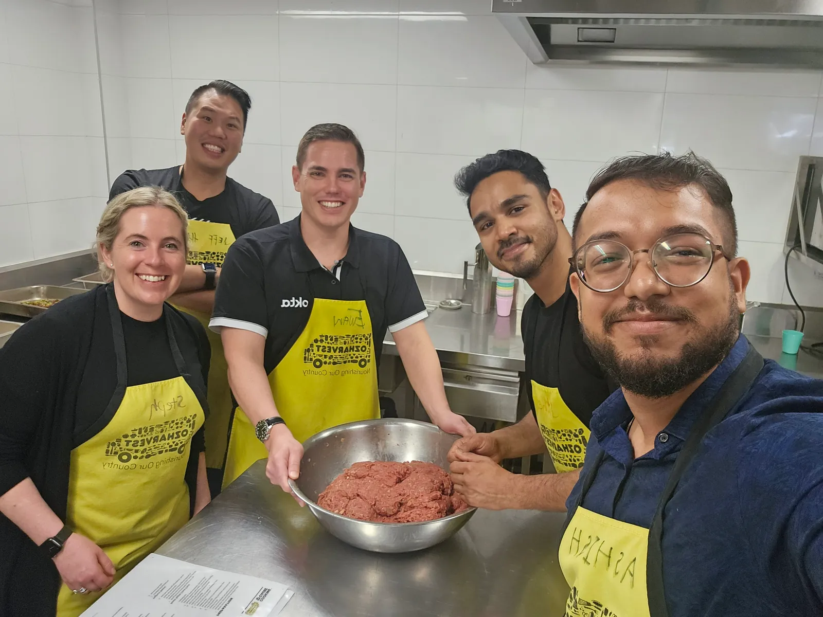 Ashish taking a group selfie with colleagues in yellow OzHarvest aprons around a mixing bowl.