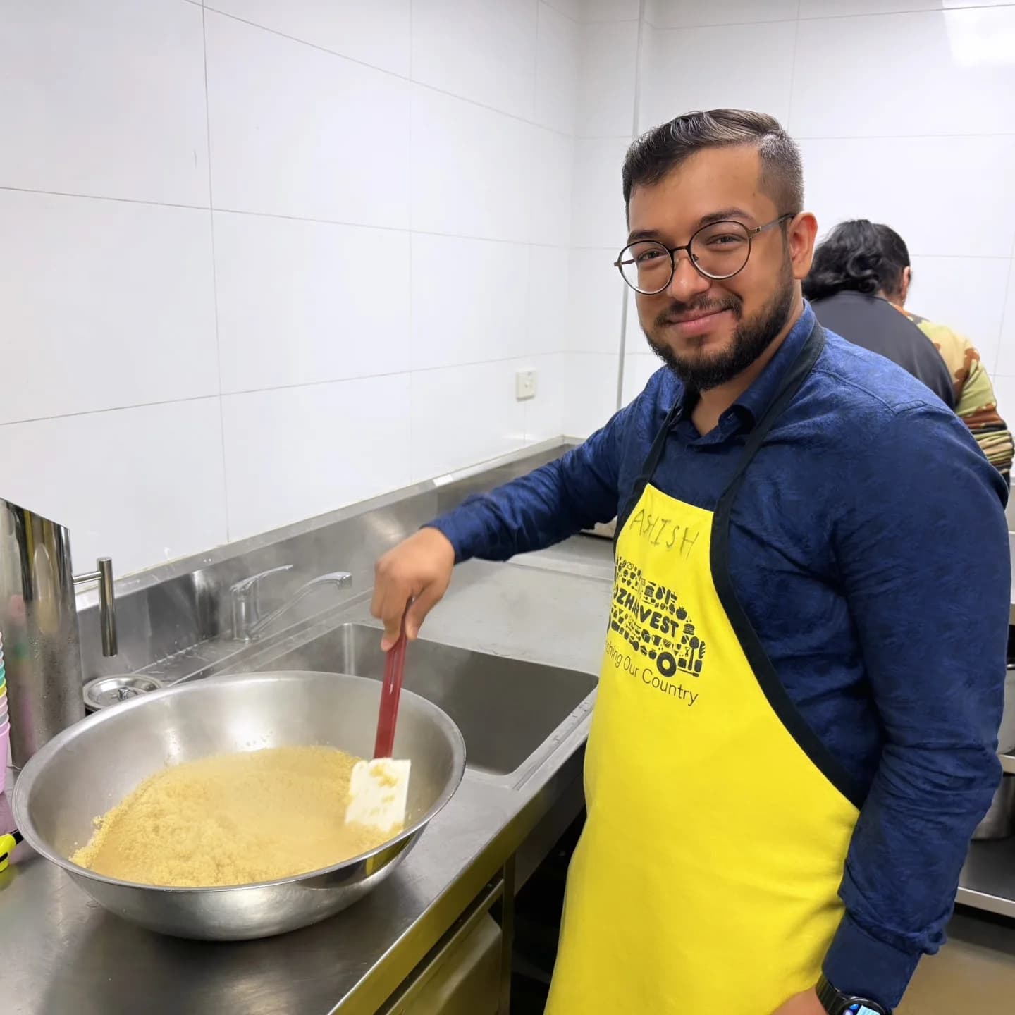 Ashish in a yellow OzHarvest apron, stirring a large pot in a kitchen.