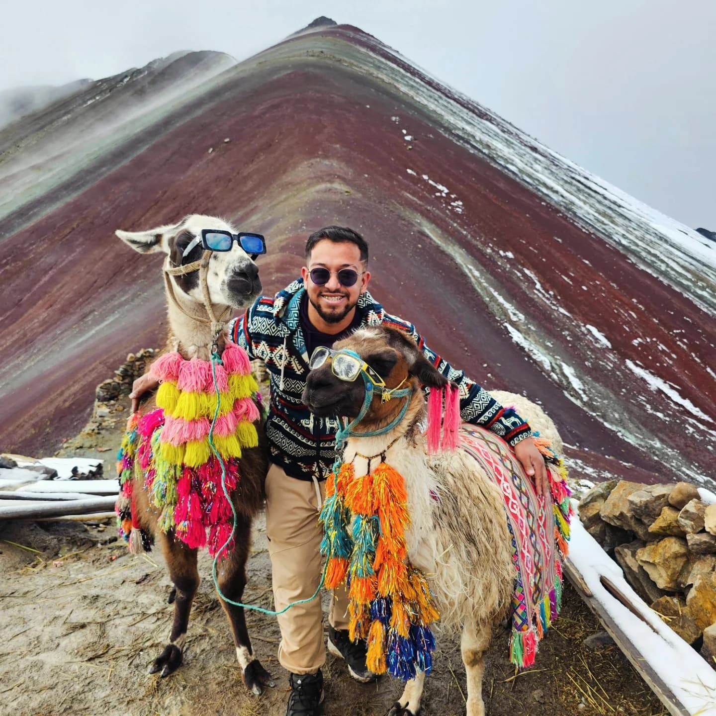 Ashish between two decorated alpacas wearing sunglasses, with the striped slopes of Rainbow Mountain behind them.