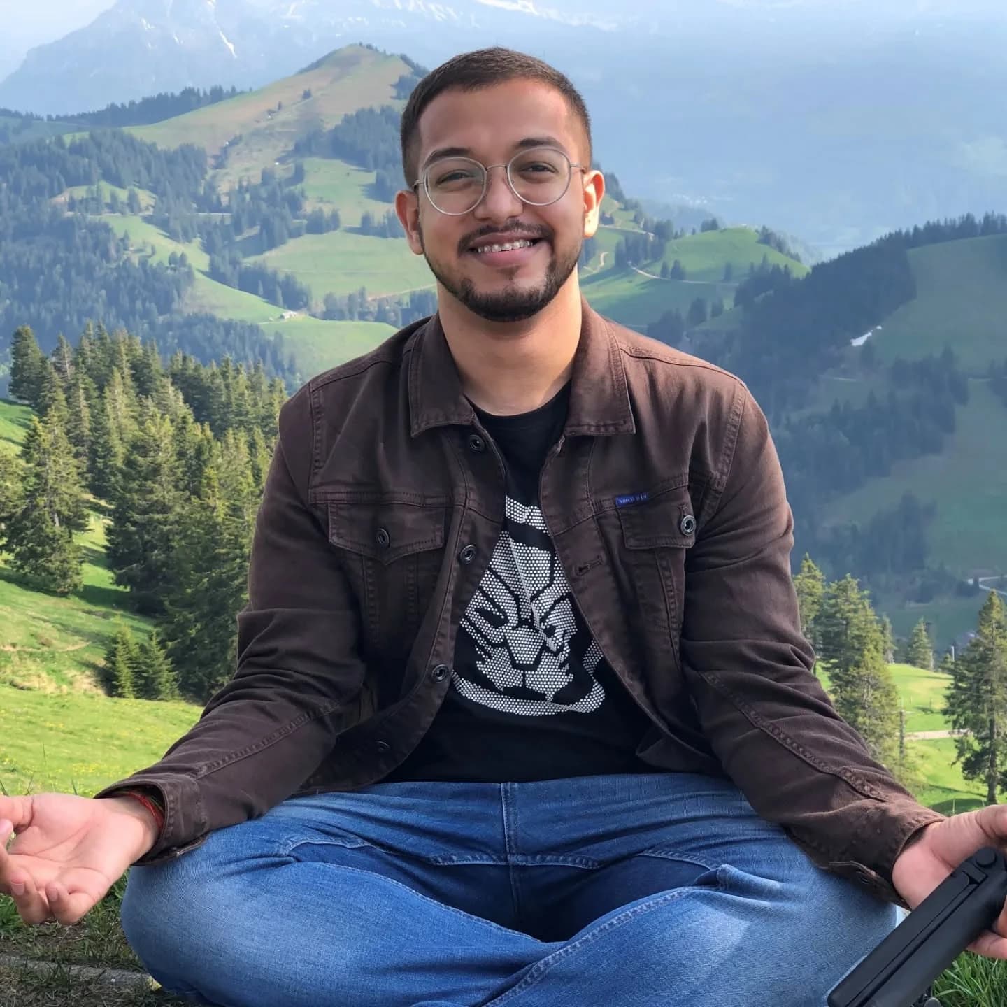 Ashish smiling and sitting cross-legged at Mount Rigi, with green hills, forests and mountains behind him.
