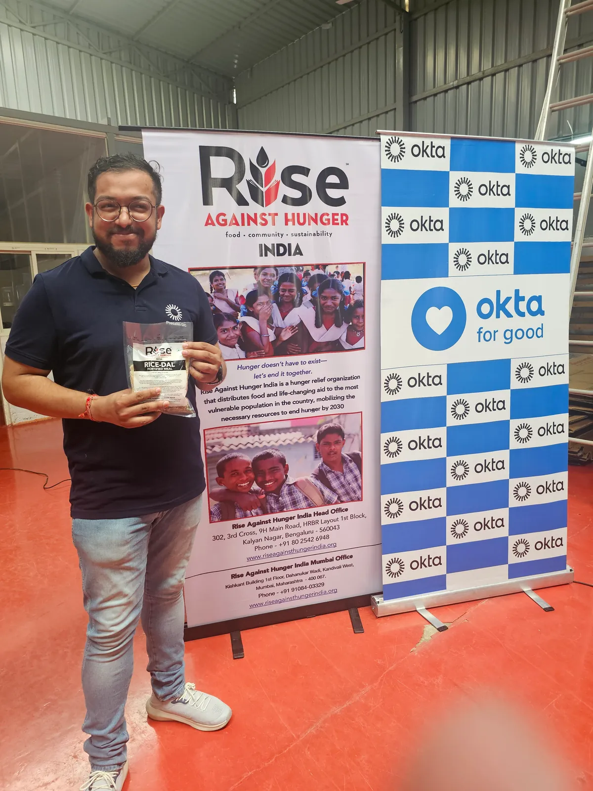 Ashish holding a packed meal beside the Rise Against Hunger and Okta for Good banners.