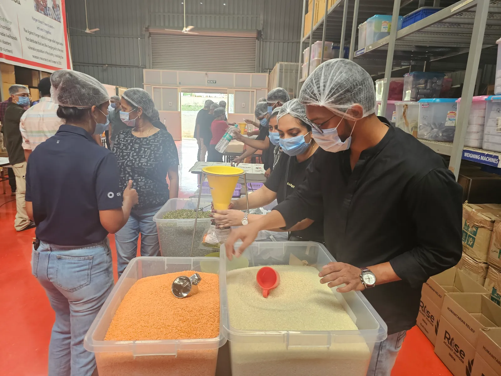 Ashish in a hairnet and mask, portioning ingredients into a packet at a meal-packing station.