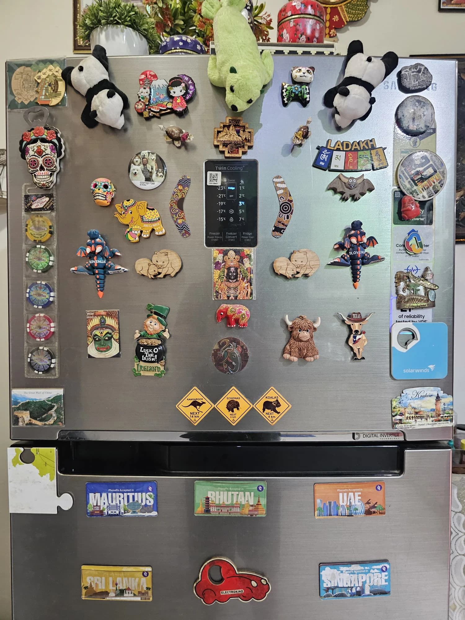 A silver fridge covered with travel souvenirs: plush pandas, a green crocodile, colourful skulls, Slovenian dragons, a leprechaun, and magnets from places including Sri Lanka, Bhutan, Singapore and Mauritius.
