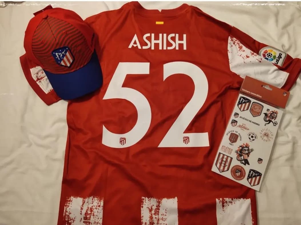 An Atlético Madrid home shirt laid flat with a matching cap and a sheet of club stickers. ASHISH is printed across the shoulders above the number 52, with the LaLiga patch on the sleeve.